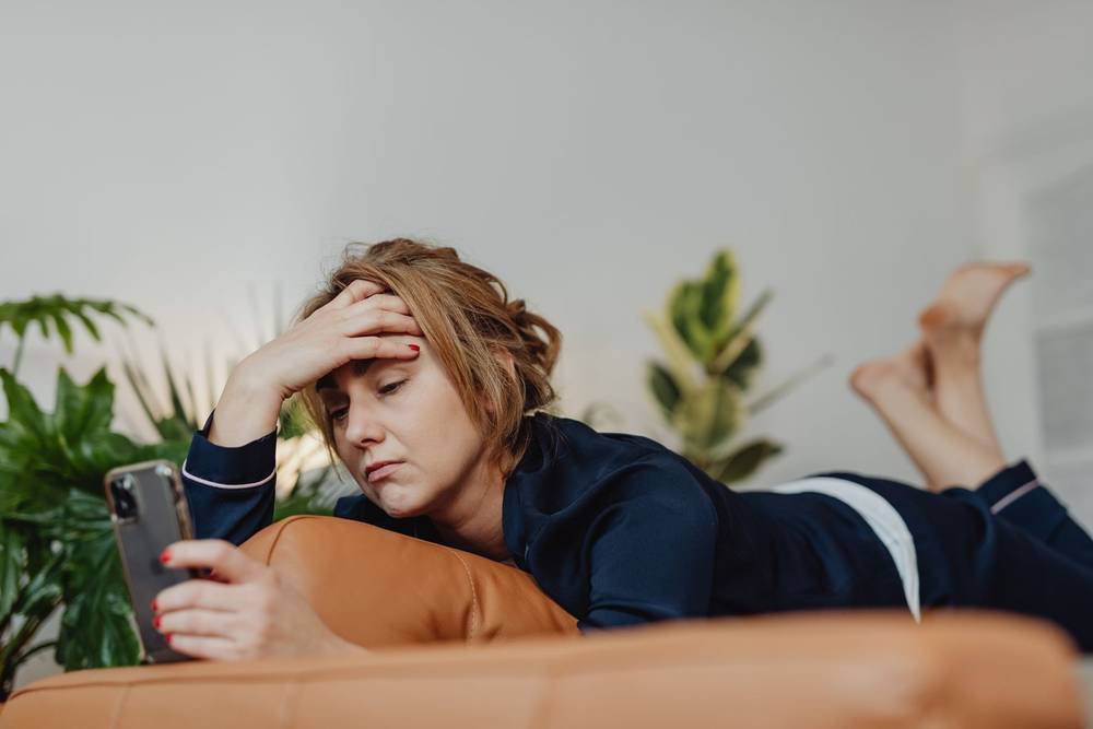 Eine junge Frau liegt auf dem Sofa, hält ihr Handy und fasst sich besorgt an die Stirn